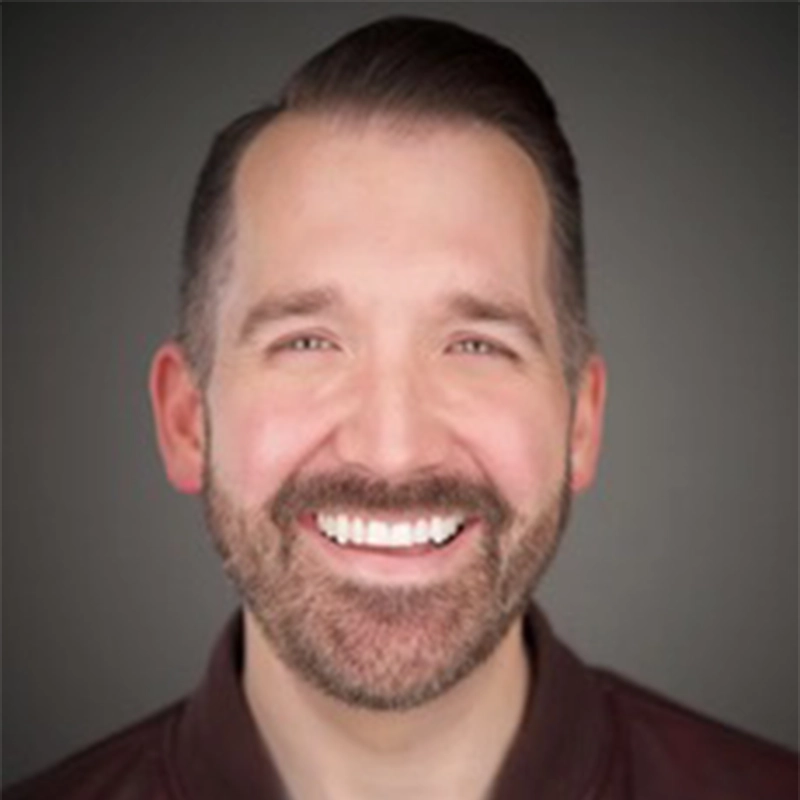 A man with fair skin, neatly styled brown hair, and a trimmed beard smiles broadly, showing his teeth. He is wearing a brown collared shirt and is photographed against a plain, dark background. The lighting is even, highlighting his face.