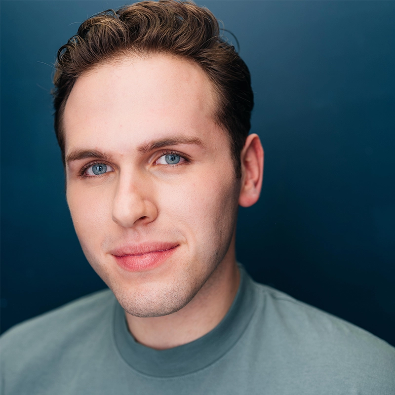 A young man with fair skin, short brown hair, and blue eyes smiles softly at the camera. He is wearing a gray crew-neck shirt and is posed in front of a solid dark blue background, reminiscent of a portrait by Sophie Smith-Brody.