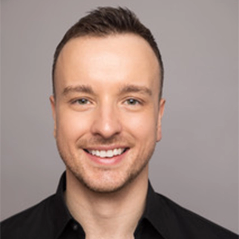 A man with short brown hair smiles at the camera. He has fair skin, hazel eyes, light facial stubble, and is wearing a black collared shirt. The background is a plain light grey. The photo is well-lit and professionally taken.