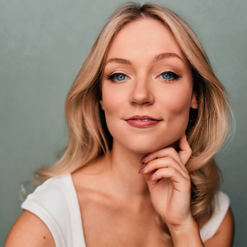 A young woman with long blonde hair and blue eyes poses against a muted green background. She wears a white top and has a gentle smile. Her right hand is lightly touching her chin, and she has natural makeup with pink lipstick and subtle eyeliner. The image captures Lucy Rhoades in a serene and graceful moment.