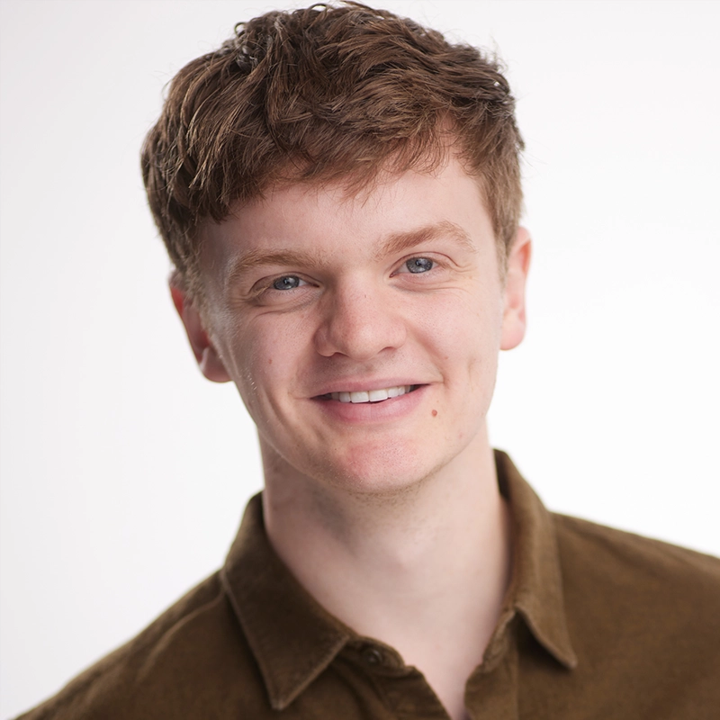 A young man with short, tousled light brown hair smiles at the camera. He wears a brown collared shirt and stands against a plain, softly lit white background. His friendly, relaxed expression makes Bailey McCall seem approachable and genuine.