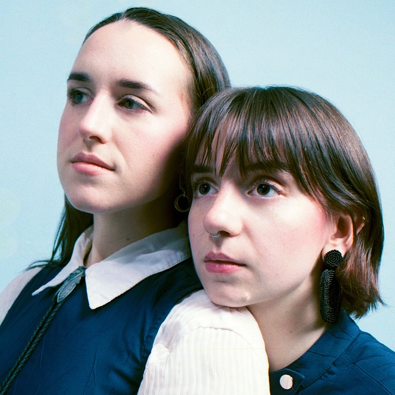 Two young women pose closely against a pale blue background. Sophie Smith-Brody stands slightly behind, resting her chin gently on the other’s head. Both wear blue vests over white shirts and have neutral expressions, gazing into the distance.