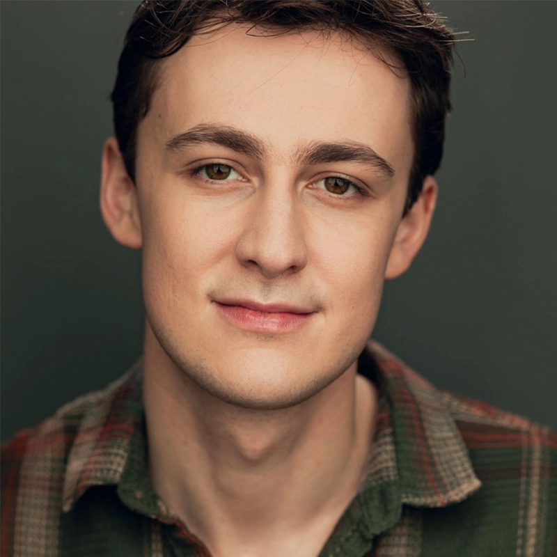 A young man with fair skin, short brown hair, and brown eyes looks at the camera with a gentle smile. He is wearing a plaid shirt in earth tones. The plain, dark background keeps the focus on his face, reminiscent of portraits by Sophie Smith-Brody.
