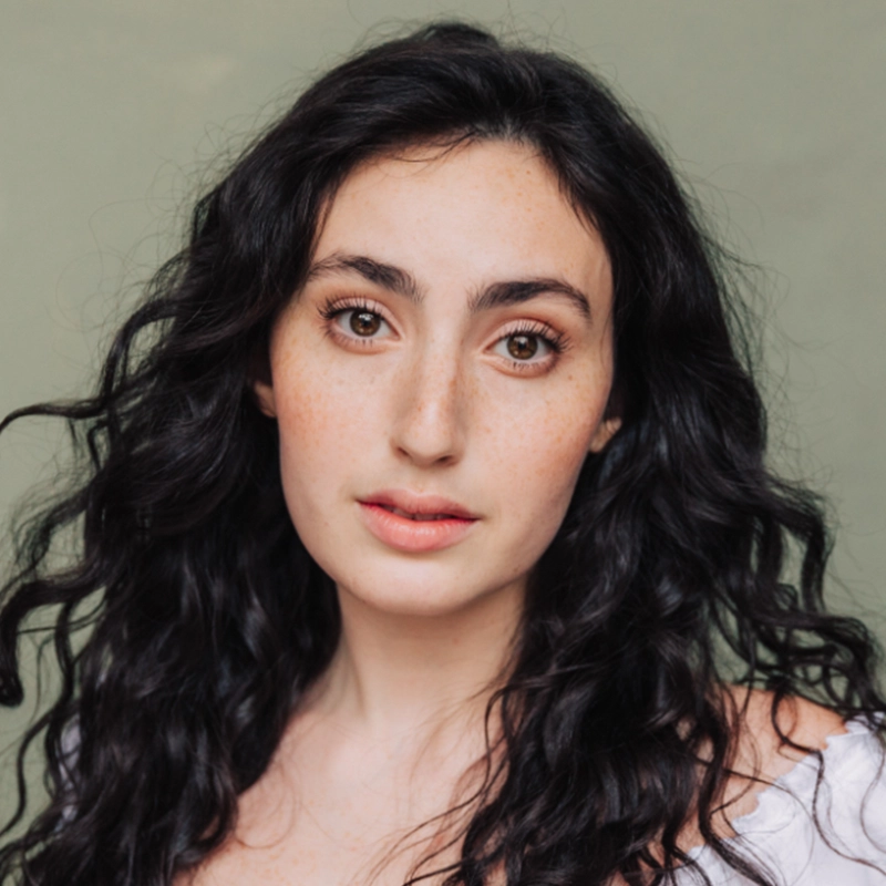 A young woman with long, wavy dark hair and fair skin looks at the camera. She has brown eyes, natural makeup, and soft pink lips. Sophie Smith-Brody wears a white top off one shoulder against a plain, muted green background.