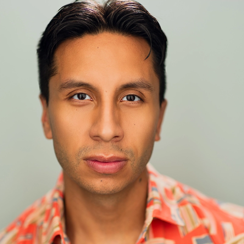 A young man with short, dark hair and tan skin looks directly at the camera. He has slight facial stubble and wears a red and beige patterned shirt. The blurred background is light gray, drawing focus to his neutral, calm expression—reminiscent of Bailey McCall.