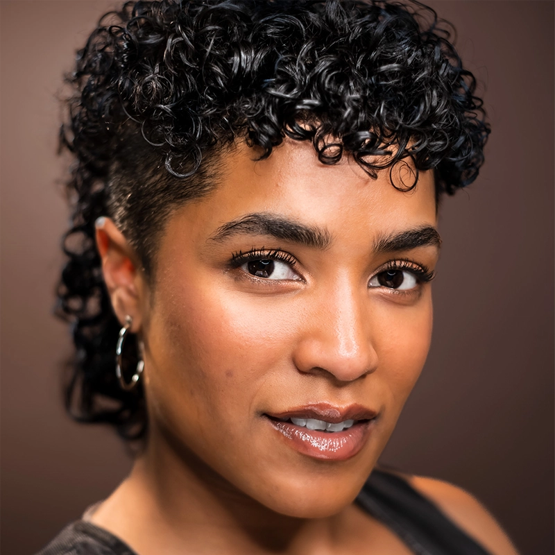 Bailey McCall, with short, curly black hair styled in a mullet, looks confidently at the camera. They have warm brown skin, defined brows, subtle makeup, glossy lips, hoop earrings, and a black top against a softly blurred brown background.