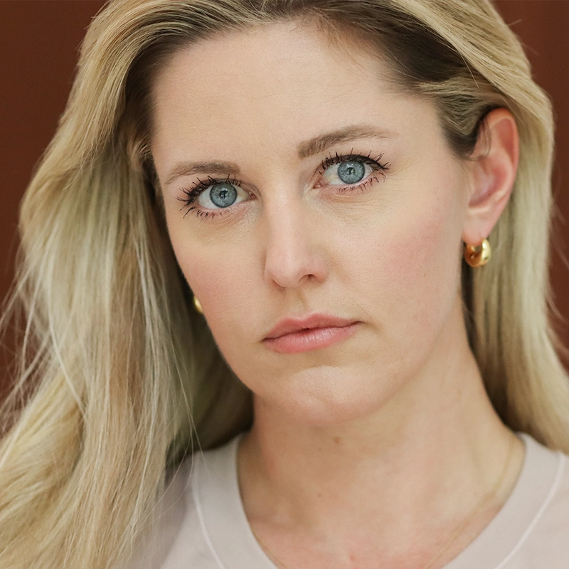 Bailey McCall, with long blonde hair and blue eyes, looks directly at the camera with a serious expression. She wears gold hoop earrings and a light-colored top against a plain, out-of-focus background that draws attention to her face.