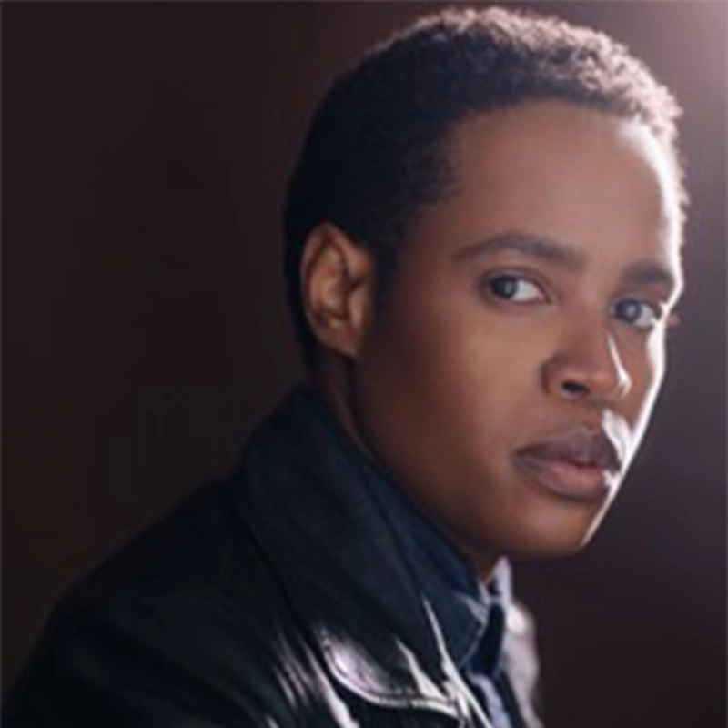 A person with short hair and medium-dark skin gazes seriously at the camera. They wear a dark jacket over a shirt. The lighting is soft and highlights one side of their face, set against a dark, blurred background.