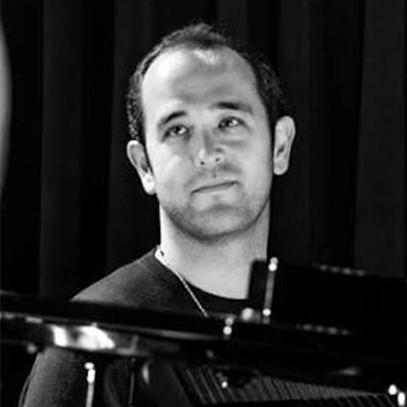 A man with short dark hair and a trimmed beard looks upward thoughtfully. He sits behind a piano, dark curtains in the background; the black and white image evokes a contemplative mood reminiscent of Sophie Smith-Brody’s introspective portraits.