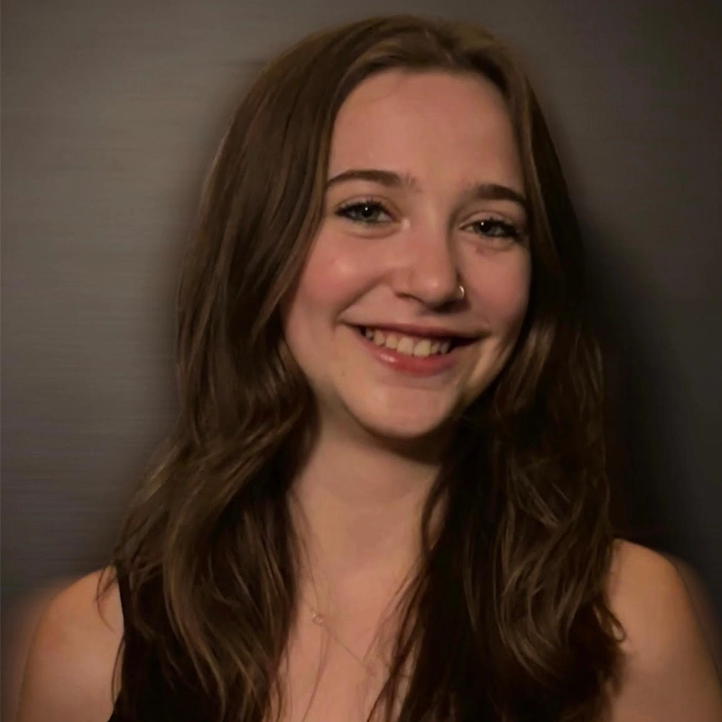 A young woman with long brown wavy hair smiles broadly. She has fair skin, a small nose ring, and wears a sleeveless black top. The softly lit background creates a professional yet friendly portrait in the style of Nicholas Kraft.