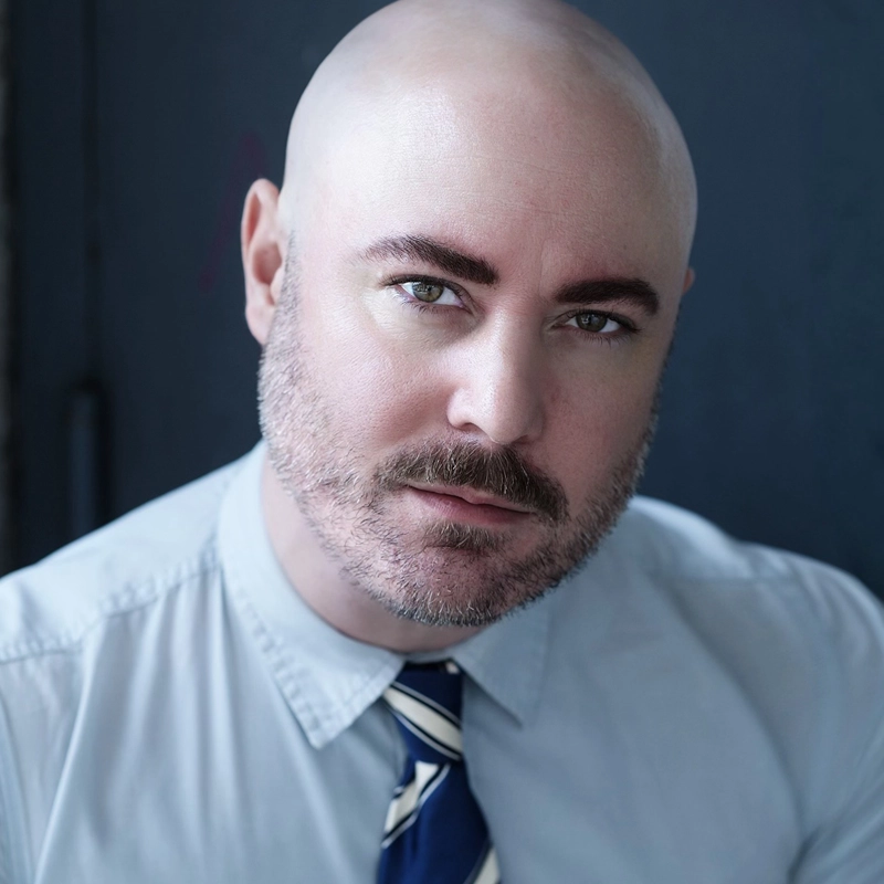 Ryan Bauer-Walsh, a bald man with a trimmed beard and mustache, gazes confidently at the camera. He sports a light blue collared shirt and a navy and white striped tie against a dark, blurred background that draws focus to his expression.