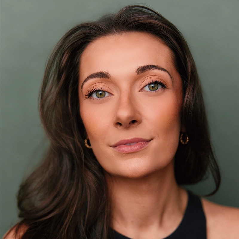 Carrie Wagner, with long brown hair and green eyes, gazes at the camera with a slight smile. She wears a dark sleeveless top and small hoop earrings against a solid, muted green background. Even lighting highlights her features.