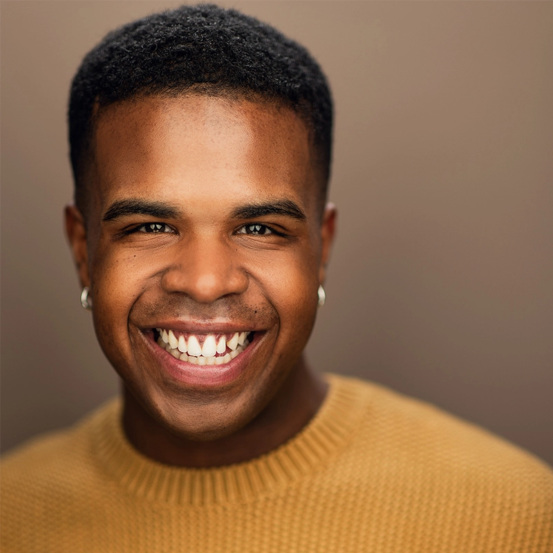 Corey Barrow, a young man with short curly hair and medium brown skin, smiles widely, showing his teeth. He wears small hoop earrings and a mustard yellow knit sweater. The softly blurred brown background highlights his cheerful expression.