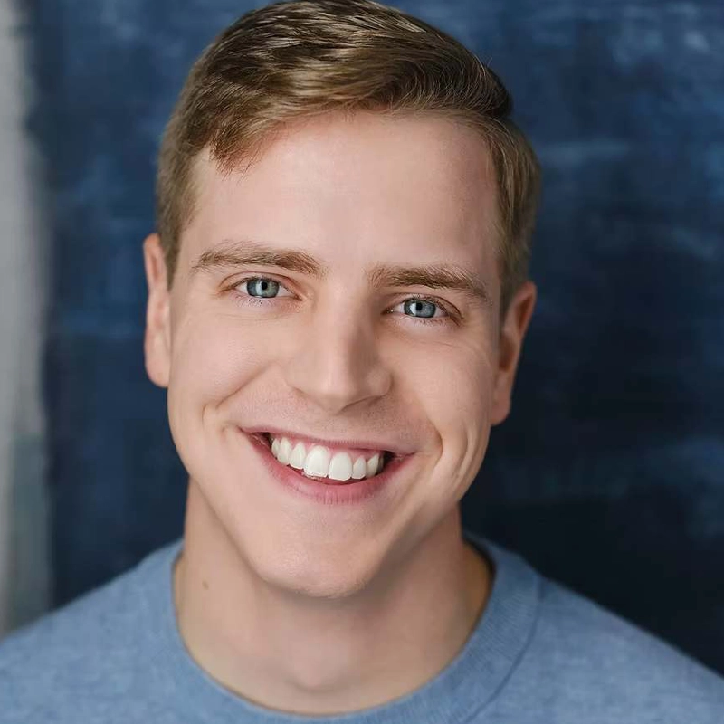 Jackson Hurt, a young man with short light brown hair and blue eyes, smiles widely, showing his teeth. He is wearing a light blue crew-neck shirt against a softly blurred dark blue and gray gradient background.
