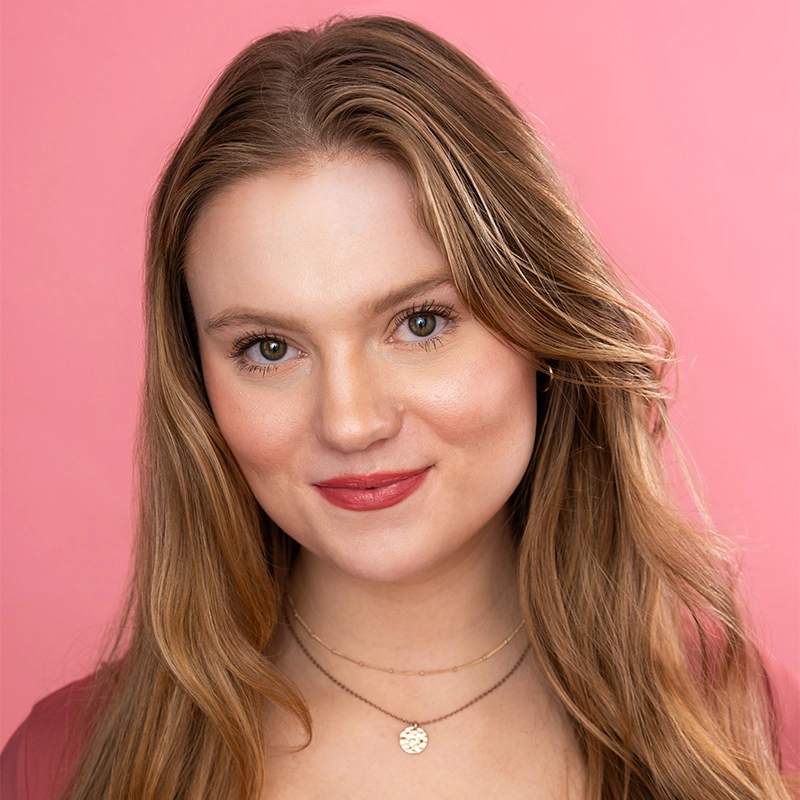 A young woman with long, light brown hair and fair skin smiles softly against a pink background. Styled by Nicholas Kraft, she wears layered gold necklaces and a pink top. Her natural makeup gives her rosy cheeks and lips as she looks directly at the camera.