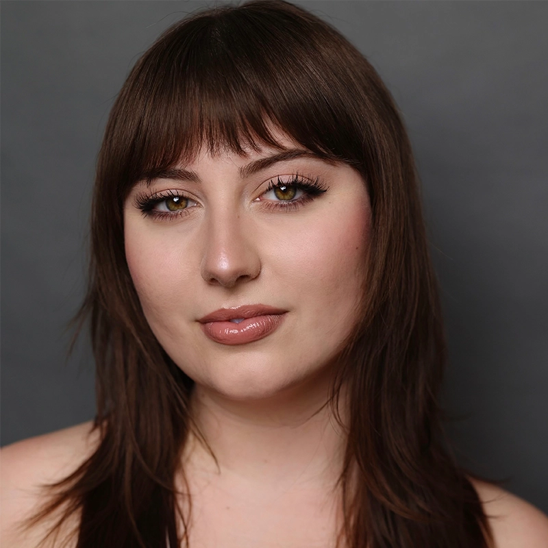 A young woman with fair skin and medium-length straight brown hair with bangs is facing the camera. She has hazel eyes, long lashes, subtle makeup, and glossy lips. Photographed in the style of Nicholas Kraft against a plain, dark gray background.