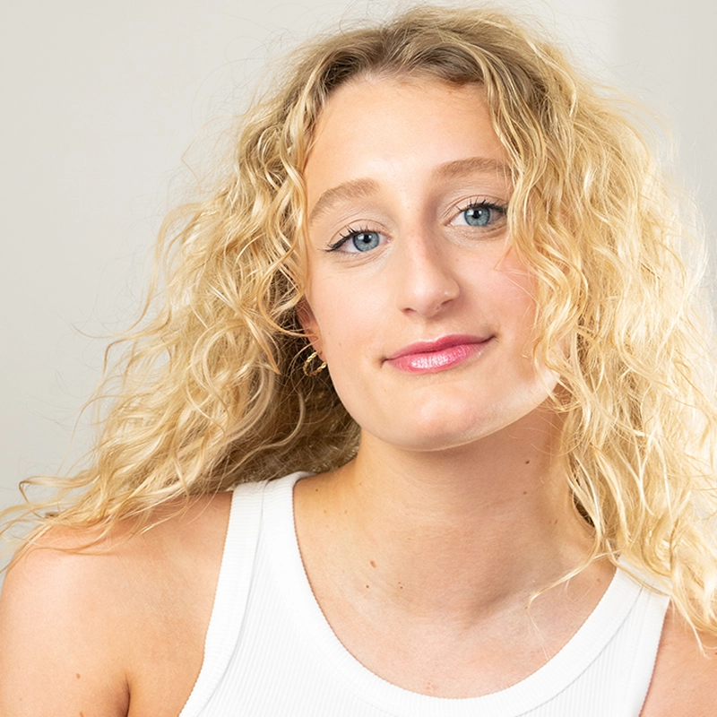 A young woman, Maddie Conti, with long, curly blonde hair and blue eyes smiles softly. She wears a white sleeveless top and minimal makeup. The background is plain and light-colored, giving the image a bright, fresh feel.