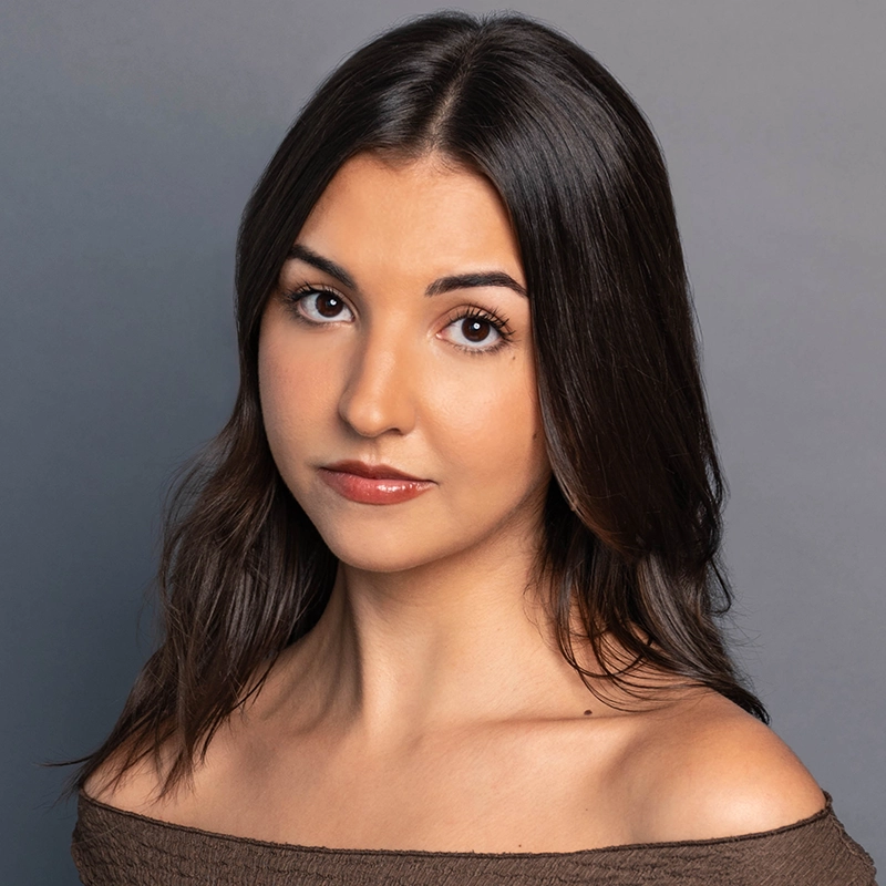 Maddie Conti, a young woman with long, dark brown hair and brown eyes, looks directly at the camera with a neutral expression. She wears an off-the-shoulder brown top against a plain gray background.