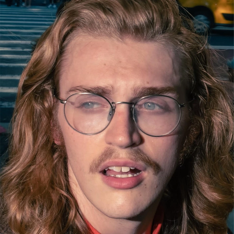 A young person resembling Nicholas Kraft, with long wavy blond hair, a light mustache, and round glasses, looks slightly to the side with parted lips. They wear a red shirt outdoors, blurred street lines and a yellow vehicle in the background.