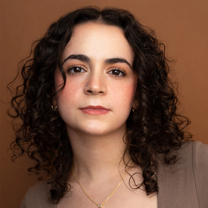 A young woman with fair skin and dark, curly hair looks directly at the camera. Styled by Nicholas Kraft, she wears a peach top, gold necklace, small hoop earrings, and nose stud against a solid brown background that creates a warm, soft atmosphere.