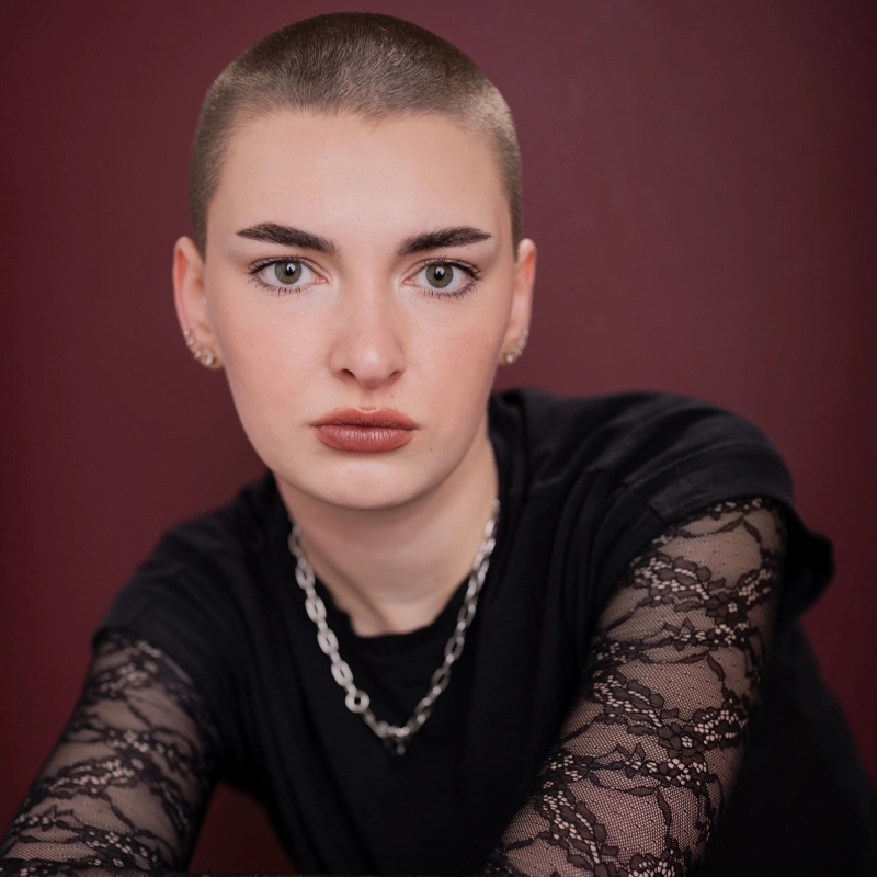Sophie Tyler, with closely shaved hair and bold eyebrows, stares directly at the camera. She wears a black top with lace sleeves, a silver chain necklace, and small earrings against a solid dark burgundy background.