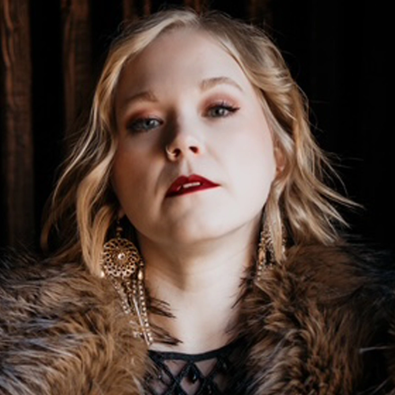 A woman with wavy blonde hair, dark red lipstick, and dramatic eye makeup looks confidently at the camera. She wears a fur-like coat, ornate gold earrings, and a black lace top, standing against a dark background with vertical wood-like textures.