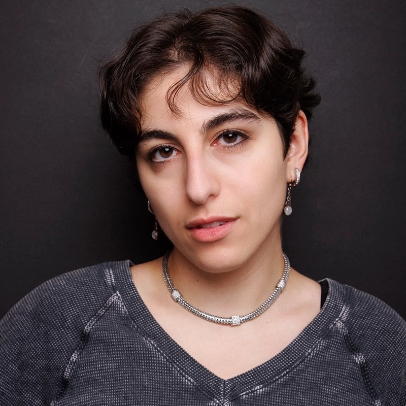 Alyssa Cassese, with short, wavy brown hair, poses against a dark background. She wears a gray textured shirt, silver necklace, and drop earrings. Her expression is neutral as she looks slightly off-camera with a relaxed posture.