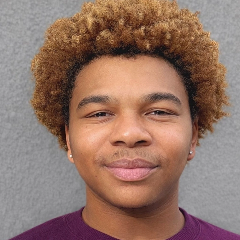 A young person with light brown skin and short curly blond hair smiles softly at the camera. They wear a purple shirt and small stud earrings, standing in front of a plain gray textured wall.