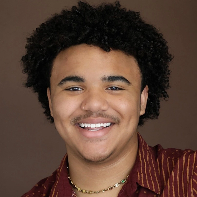 A young person with short curly hair and a nose ring smiles at the camera. They wear a maroon shirt with narrow yellow stripes and a gold chain necklace. The background is a plain, warm brown color.