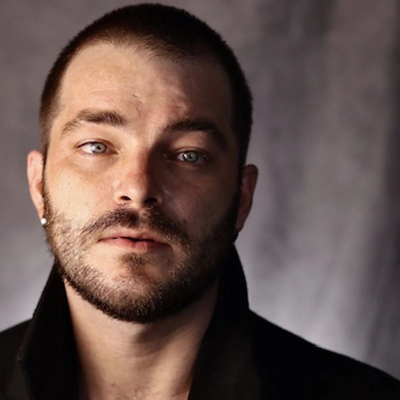 Randall Scott Carpenter, a man with short brown hair, a trimmed beard, and light blue eyes, stares at the camera. He wears a black coat with the collar up against a softly blurred gray background. His expression is serious and contemplative.