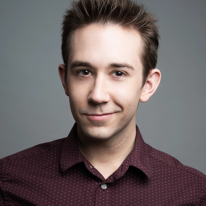 Jackson Cline, a young man with short, spiked brown hair, smiles softly at the camera. He wears a maroon button-up shirt with small white polka dots against a plain gray background, lit with soft and even lighting.