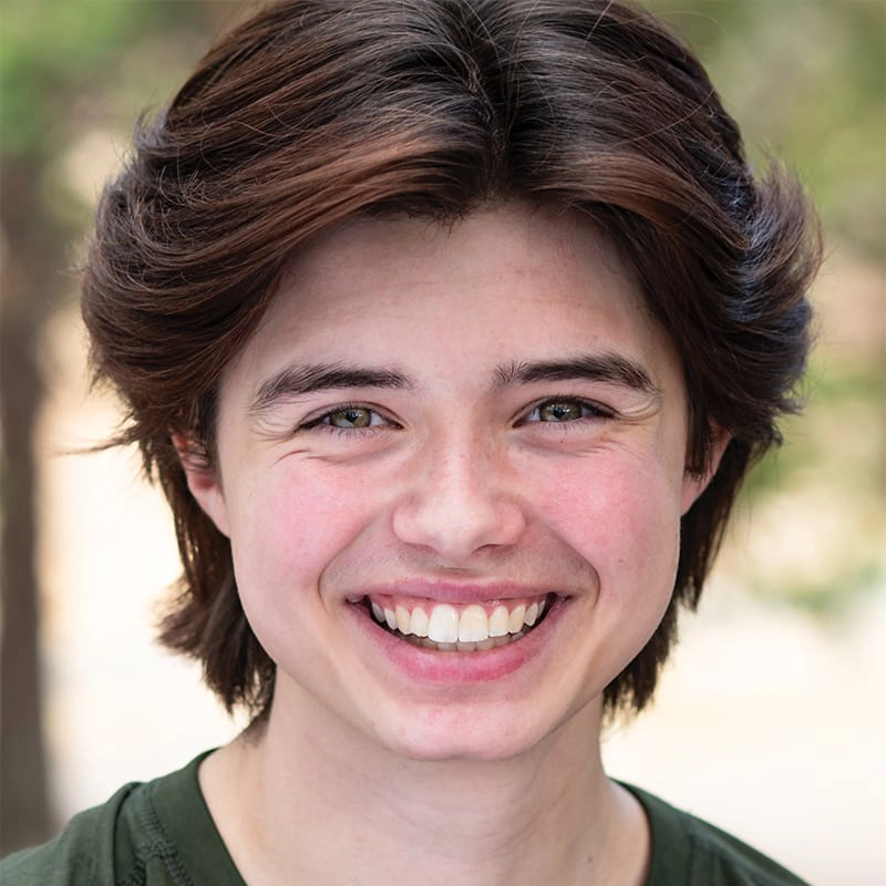A young person with medium-length brown hair smiles widely, showing teeth. They have light skin, green eyes, and are wearing a dark green shirt. The background is softly blurred with hints of greenery, suggesting an outdoor setting.