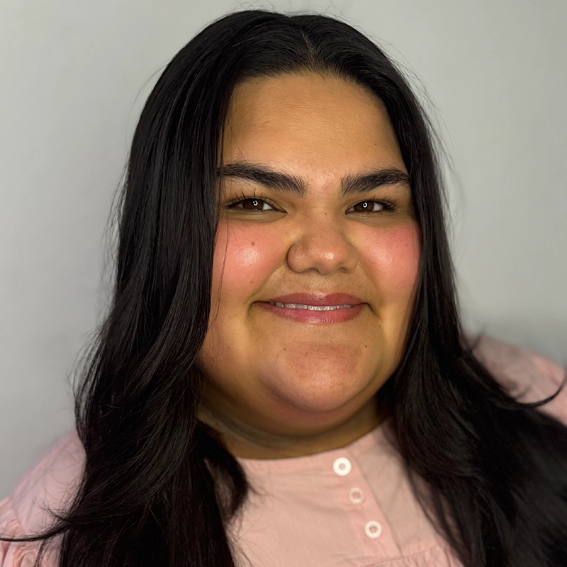 A woman with long dark hair smiles warmly at the camera. She has full cheeks, smooth skin, and rosy blush. She is wearing a light pink blouse with buttons and is posed in front of a plain light gray background.