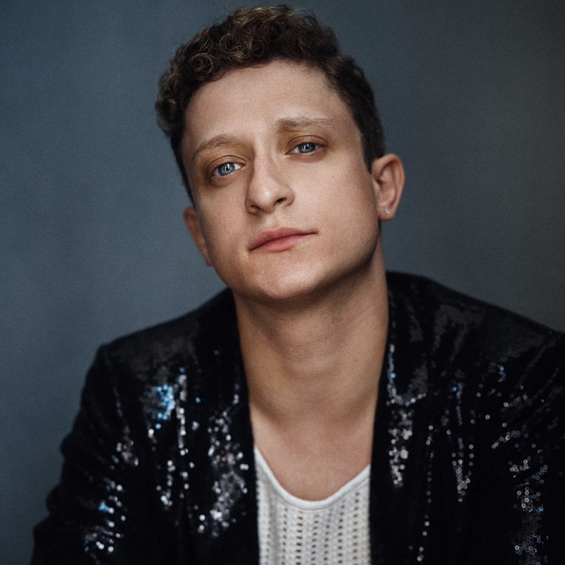 Dylan Hoffinger, with short curly hair and blue eyes, looks slightly up at the camera, wearing a black sequin jacket over a white knit top against a plain dark background. Their expression is neutral and composed.