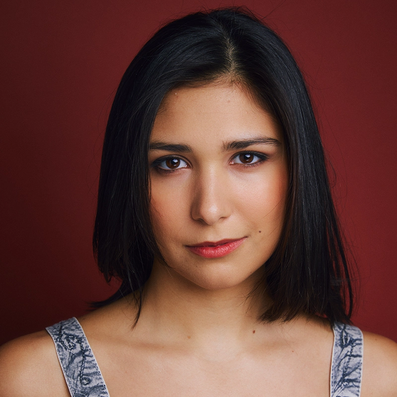 A young woman with straight, shoulder-length dark hair is looking directly at the camera. She wears a patterned tank top and subtle makeup. The background is solid maroon, and the lighting highlights her face. Her expression is calm and neutral.