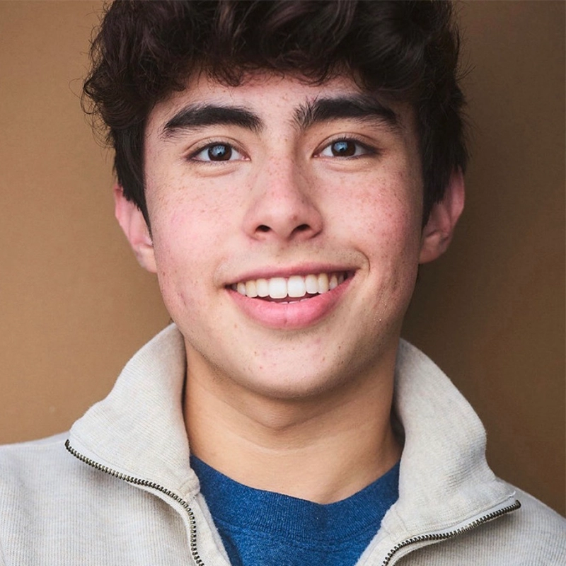 A young person with short, curly dark hair smiles at the camera. They have clear skin with faint freckles, thick eyebrows, and are wearing a cream-colored zip-up jacket over a blue shirt. The background is plain and brown.
