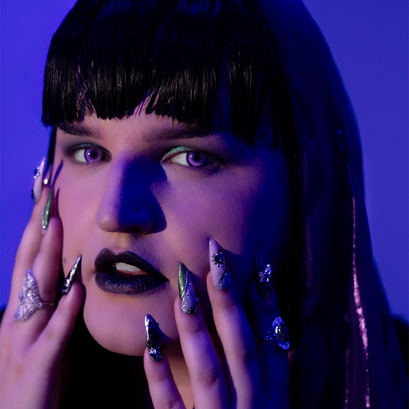 Person with dramatic makeup, dark lipstick, green eyeshadow, and long, decorated nails poses with fingers near face. Wearing a dark veil and multiple rings, they stare intensely. Lighting is blue-purple, creating a bold, mysterious atmosphere.