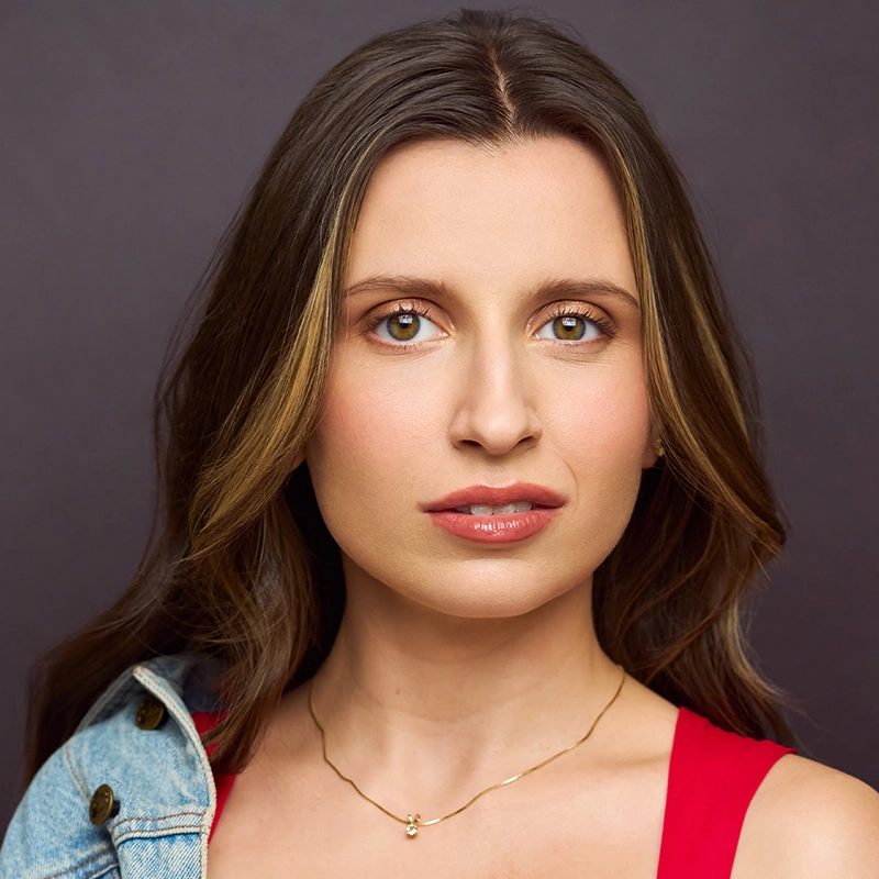 A woman with long brown hair and highlighted strands stares directly ahead. She wears a red top, a light denim jacket over one shoulder, and a delicate gold necklace. The background is plain and dark, emphasizing her serious expression.