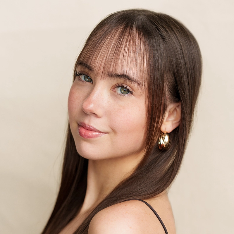 A young woman with long, straight brown hair and wispy bangs smiles softly. She wears gold hoop earrings and a black spaghetti strap top, with light pink lipstick and natural makeup. The background is a plain, light beige color.