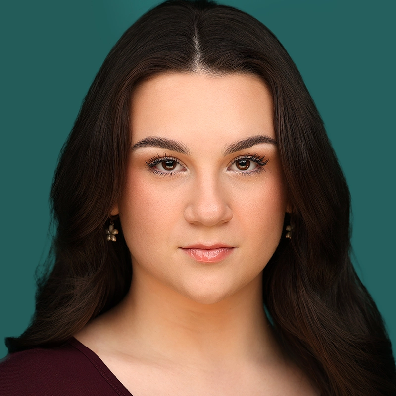 A young woman with long, dark brown hair and fair skin looks directly at the camera, wearing natural makeup and gold earrings. She is dressed in a burgundy top, and the background is a solid teal color.