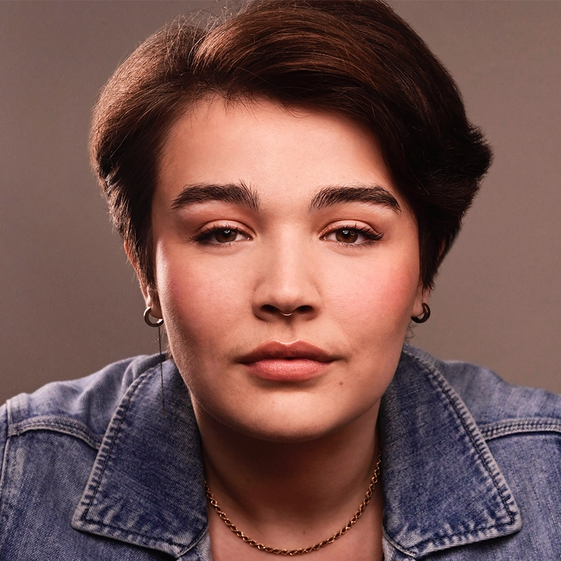 Kat Rodriguez, with short brown hair, light skin, and natural makeup, looks directly at the camera. She wears a denim jacket, a gold chain necklace, and small hoop earrings against a plain, softly lit background.
