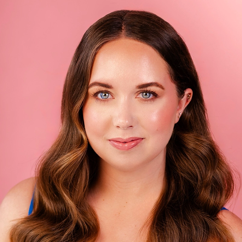 A woman with long, wavy brown hair and blue eyes looks at the camera, smiling softly. She has glowing, natural makeup and wears a blue top. The background is a soft pink color, creating a warm and inviting atmosphere.