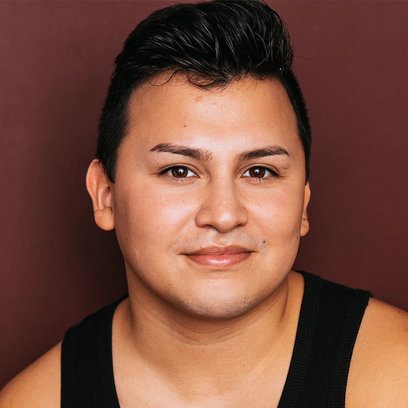A person with short dark hair and smooth skin smiles gently. They wear a black tank top and pose in front of a plain maroon background. The lighting is soft, highlighting their facial features and creating a warm, approachable atmosphere.