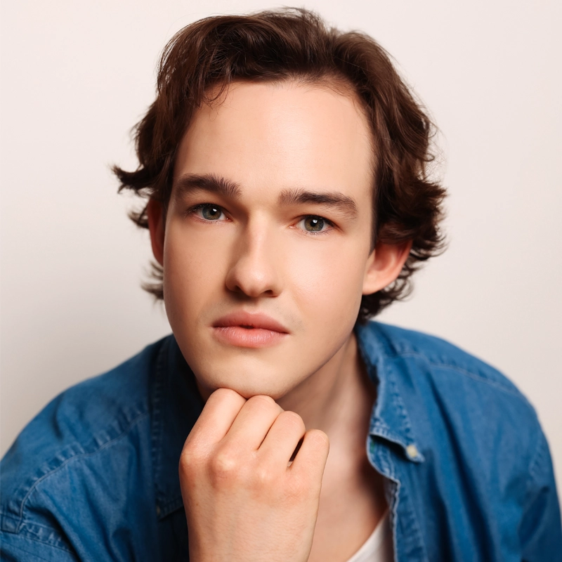 A young person with fair skin, short brown wavy hair, and blue eyes gazes softly at the camera. Andrew Tompkins Lewis rests their chin on their hand, wearing a blue denim shirt over a white top against a neutral background.