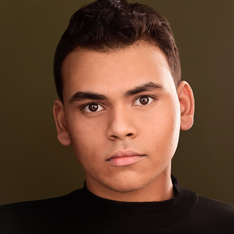 A young man with short, dark curly hair and brown eyes stares directly at the camera. He has a neutral expression and wears a black turtleneck. The background is a muted olive green, and the lighting is soft, highlighting his facial features.