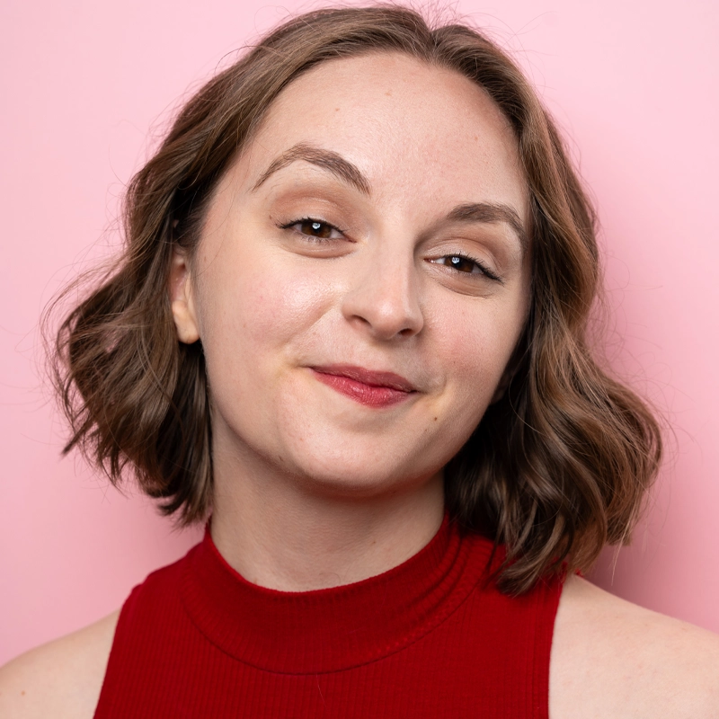 Lora Margerum, a young woman with wavy brown hair and fair skin, smiles softly. She wears a sleeveless red top and light makeup. The solid pink background adds a cheerful, warm tone to the portrait.