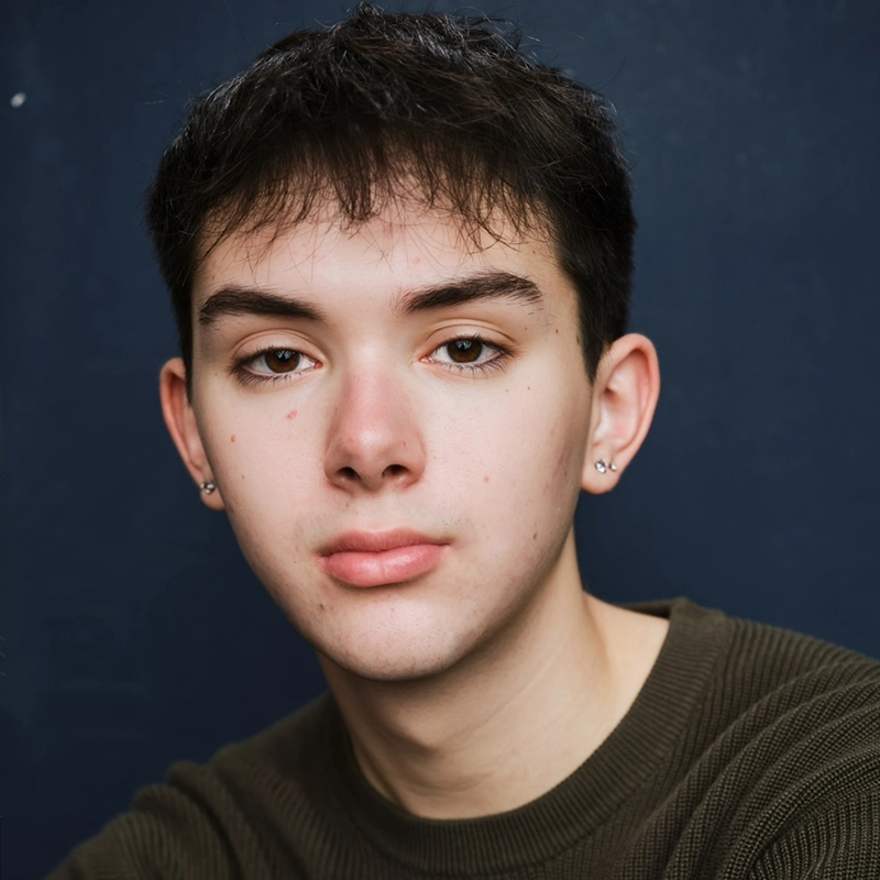 Young person with short dark hair, fair skin, and brown eyes looks directly at the camera. They wear a dark green ribbed sweater and small stud earrings in both ears. The background is a plain, dark blue color. Their expression is neutral and calm.