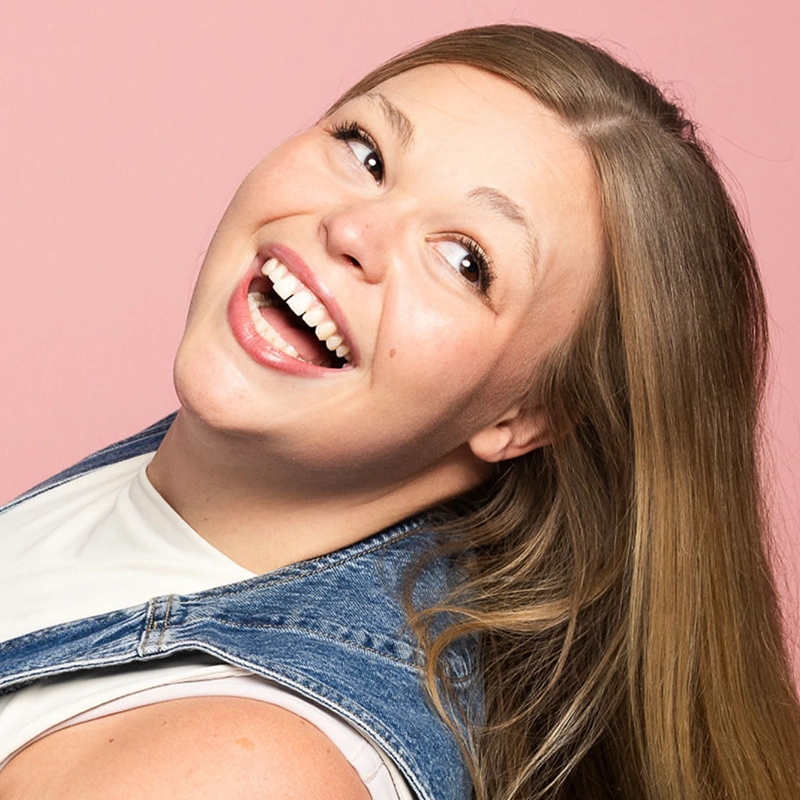 A smiling woman with long, straight blonde hair poses with her head tilted back. She wears a white top under a blue denim vest. The background is pale pink, and she looks happy and cheerful with her mouth open and eyes wide.