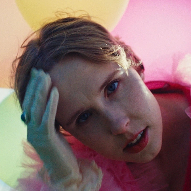 Kat Rodriguez, with short light brown hair and blue eyes, gazes at the camera, tilting their head and touching their forehead with a gloved hand. They wear a pink, feathery garment against soft-focus pastel yellow and pink balloons.