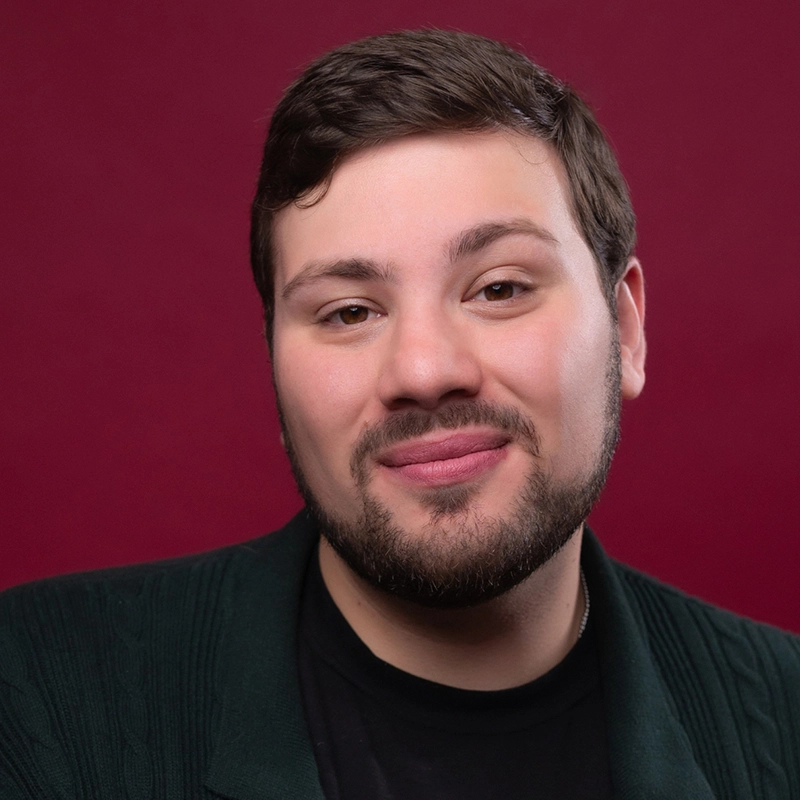 A person with short dark hair and a trimmed beard smiles gently at the camera. They wear a dark top and a green textured sweater. The background is solid burgundy. The lighting is soft, giving the image a warm, welcoming feel.